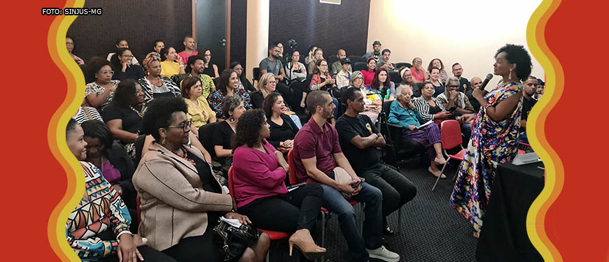 Visão do auditório lotado durante a palestra da Dra. Lívia Sant'Anna Vaz, que fala ao microfone. Ela é uma mulher negra de cabelos cacheados de comprimento médio, ela veste um vestido com estampa colorida.