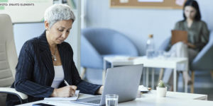 Mulher com cabelos cabelos curtos e grisalhos sentada em uma mesa diante de um computador em um escritório. Ela está com uma expressão séria e concentrada. Ao fundo está uma colega de trabalho.