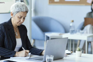 Mulher com cabelos cabelos curtos e grisalhos sentada em uma mesa diante de um computador em um escritório. Ela está com uma expressão séria e concentrada. Ao fundo está uma colega de trabalho.