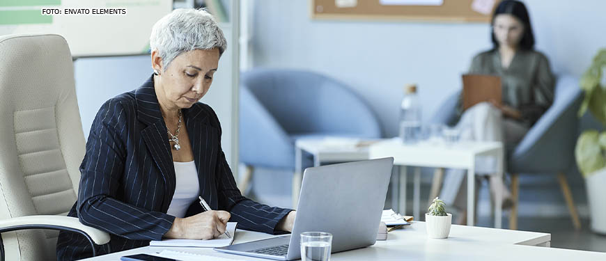 Mulher com cabelos cabelos curtos e grisalhos sentada em uma mesa diante de um computador em um escritório. Ela está com uma expressão séria e concentrada. Ao fundo está uma colega de trabalho.