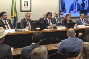 A imagem mostra uma audiência pública no plenário de uma comissão parlamentar, com a presença de parlamentares e representantes de entidades.