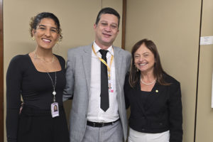 Três pessoas posam sorrindo para a foto em um ambiente interno. À esquerda, uma mulher de cabelo cacheado preso, usando blusa preta e crachá. Ao centro, um homem de terno cinza, camisa branca e gravata preta. À direita, uma mulher de cabelo liso castanho, vestindo blazer preto e calça branca.