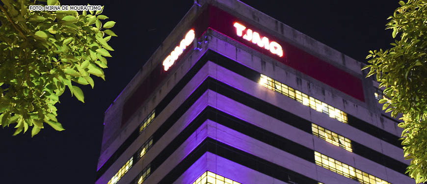Vista noturna da fachada da sede do TJMG iluminada com luzes rosadas. Conteúdo textual: TJMG divulga intimação de candidatos à PV 2021 com documentos incorretos.