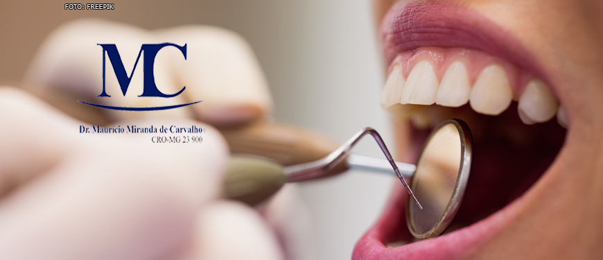 Em destaque, mão de dentista durante atendimento segurando um espelho e uma pinça perto de uma boca aberta de paciente. Na imagem tem a logo do dentista Maurício Miranda de Carvalho (CRO-MG 23.900).