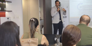 Um homem (Tarciso Palma) fala ao microfone em uma sala de palestras diante de um telão com apresentação. Há algumas pessoas sentadas assistindo. Conteúdo textual: Qualidade de vida - Fórum Permanente do SINJUS debate jornada de 8 horas e saúde no trabalho.