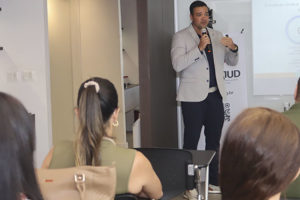 Um homem (Tarciso Palma) fala ao microfone em uma sala de palestras diante de um telão com apresentação. Há algumas pessoas sentadas assistindo. Conteúdo textual: Qualidade de vida - Fórum Permanente do SINJUS debate jornada de 8 horas e saúde no trabalho.