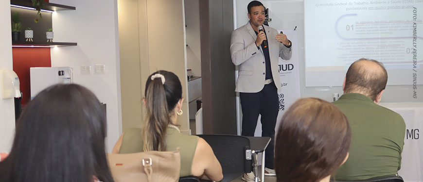 Um homem (Tarciso Palma) fala ao microfone em uma sala de palestras diante de um telão com apresentação. Há algumas pessoas sentadas assistindo. Conteúdo textual: Qualidade de vida - Fórum Permanente do SINJUS debate jornada de 8 horas e saúde no trabalho.