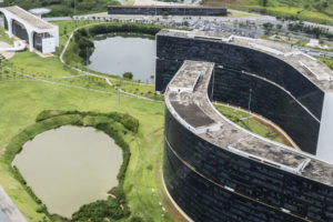 vista área da Cidade Administrativa de Minas Gerais. Em primeiro plano vemos os dois principais edifícios projetados por Oscar Niemeyer, dois lagos ornamentais. Ao fundo vemos uma paisagem de uma serra e a cidade de Belo Horizonte.