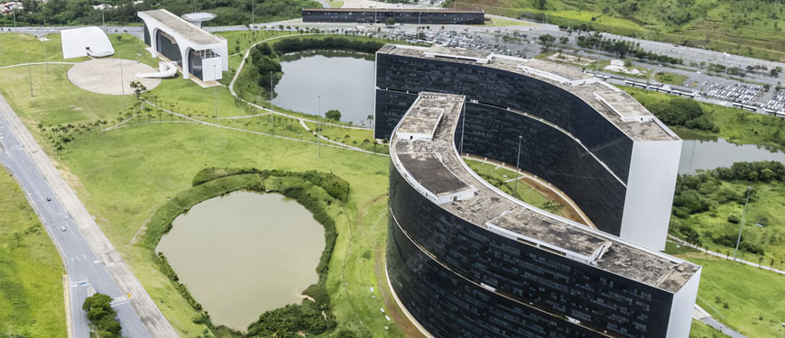 vista área da Cidade Administrativa de Minas Gerais. Em primeiro plano vemos os dois principais edifícios projetados por Oscar Niemeyer, dois lagos ornamentais. Ao fundo vemos uma paisagem de uma serra e a cidade de Belo Horizonte.