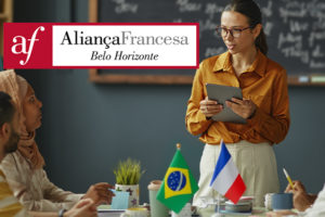 A imagem mostra uma sala de aula com uma professora segurando um tablet e sorrindo enquanto conversa com alunos sentados à mesa. Sobre a mesa, há bandeirinhas do Brasil e da França, cadernos e xícaras. Ao fundo, um quadro-negro com anotações em francês. O texto na imagem diz: “Aprenda francês com condições especiais com o novo convênio do SINJUS”. Abaixo, o logotipo da Aliança Francesa de Belo Horizonte e a marca do SINJUS-MG.