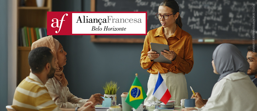 A imagem mostra uma sala de aula com uma professora segurando um tablet e sorrindo enquanto conversa com alunos sentados à mesa. Sobre a mesa, há bandeirinhas do Brasil e da França, cadernos e xícaras. Ao fundo, um quadro-negro com anotações em francês. O texto na imagem diz: “Aprenda francês com condições especiais com o novo convênio do SINJUS”. Abaixo, o logotipo da Aliança Francesa de Belo Horizonte e a marca do SINJUS-MG.