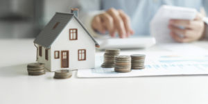 Imagem Acessível: A imagem mostra uma pessoa sentada a uma mesa enquanto confere documentos e utiliza uma calculadora, sugerindo a realização de cálculos financeiros. Em primeiro plano, há uma miniatura de uma casa e várias pilhas de moedas organizadas sobre uma folha com gráficos, simbolizando patrimônio e finanças pessoais. O ambiente tem iluminação suave e aspecto profissional. Sobre a imagem, o texto diz: “Servidores devem entregar a Declaração de Bens e Valores pelo Portal RH”. No canto superior esquerdo, lê-se “Obrigação anual”, e na base a logo do SINJUS-MG.