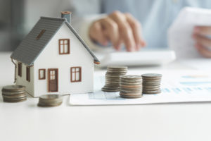 Imagem Acessível: A imagem mostra uma pessoa sentada a uma mesa enquanto confere documentos e utiliza uma calculadora, sugerindo a realização de cálculos financeiros. Em primeiro plano, há uma miniatura de uma casa e várias pilhas de moedas organizadas sobre uma folha com gráficos, simbolizando patrimônio e finanças pessoais. O ambiente tem iluminação suave e aspecto profissional. Sobre a imagem, o texto diz: “Servidores devem entregar a Declaração de Bens e Valores pelo Portal RH”. No canto superior esquerdo, lê-se “Obrigação anual”, e na base a logo do SINJUS-MG.