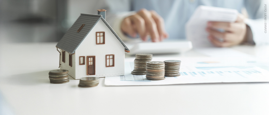 Imagem Acessível: A imagem mostra uma pessoa sentada a uma mesa enquanto confere documentos e utiliza uma calculadora, sugerindo a realização de cálculos financeiros. Em primeiro plano, há uma miniatura de uma casa e várias pilhas de moedas organizadas sobre uma folha com gráficos, simbolizando patrimônio e finanças pessoais. O ambiente tem iluminação suave e aspecto profissional. Sobre a imagem, o texto diz: “Servidores devem entregar a Declaração de Bens e Valores pelo Portal RH”. No canto superior esquerdo, lê-se “Obrigação anual”, e na base a logo do SINJUS-MG.