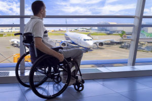 Um homem em uma cadeira de rodas está em um aeroporto, observando através de uma grande parede de vidro um avião estacionado na pista. Ele veste uma camisa clara e calça bege. O céu está azul com algumas nuvens, e a estrutura do aeroporto é moderna, com amplas janelas que permitem a visão do lado externo, onde há mais aviões e equipamentos de solo.