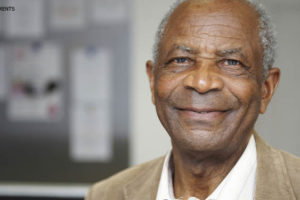 Homem negro de cabelos brancos crespos e curtos. Ele está sorrindo e veste um casaco de veludo cotelê bege por cima de uma camisa social branca.