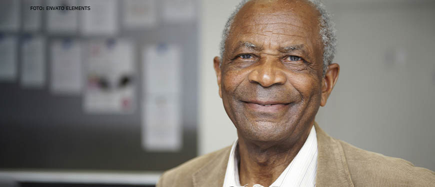 Homem negro de cabelos brancos crespos e curtos. Ele está sorrindo e veste um casaco de veludo cotelê bege por cima de uma camisa social branca.