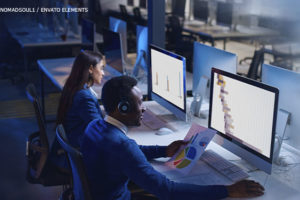 Imagem Acessível: Há duas pessoas, um homem negro e uma mulher branca em trajes sociais em frente ao computador. O ambiente está escuro, iluminado somente pelo monitor, há vários computadores em bancadas com cadeiras vazias em frente. Conteúdo textual: Plantão de habeas corpus - TJMG realiza pagamentos apenas para plantões realizados até 2023 e sindicatos criticam falta de transparência.