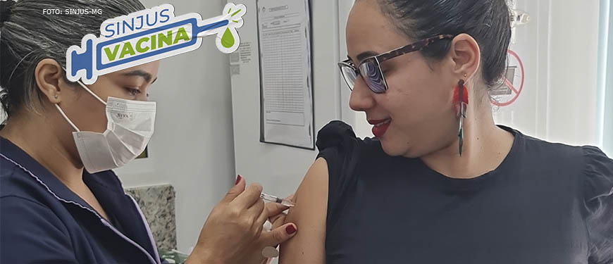 Uma mulher vestida com o uniforme de uma clínica está aplicando uma vacina no braço de outra mulher que observa o ato e sorri.