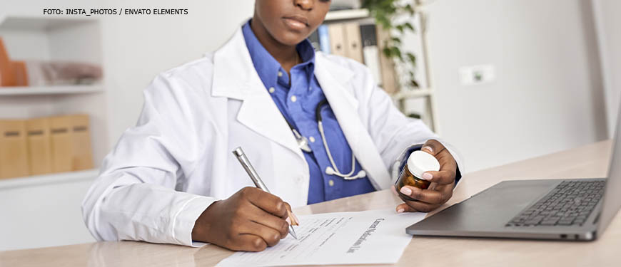 Imagem Acessível: Médica redigindo uma receita para paciente, à frente dela está um computador, em uma das suas mãos há uma embalagem de medicamento.