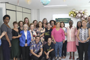 Foto das pessoas presentes na confraternização do Núcleo dos aposentados e pensionistas aplicado em cima de um fundo verde com decorações de natal.