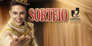 Imagem Acessível: A imagem mostra um artista em traje teatral dourado e ornamentado, estendendo uma das mãos em um gesto expressivo, sob iluminação quente e cênica. Ao fundo, há cortinas de palco desfocadas, sugerindo o ambiente de um teatro. No canto superior direito, está o logotipo da “51ª Campanha de Popularização do Teatro & Dança de Belo Horizonte”. O texto central destaca: “SORTEIO – 20 vouchers”, seguido das informações: “Período de inscrições: 13/1 (3ªf.) a 16/1 (6ªf.)” e “Resultado: 19/1 (2ªf.)”. Na parte inferior, aparece o logotipo do SINJUS-MG.