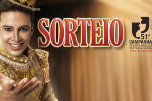 Imagem Acessível: A imagem mostra um artista em traje teatral dourado e ornamentado, estendendo uma das mãos em um gesto expressivo, sob iluminação quente e cênica. Ao fundo, há cortinas de palco desfocadas, sugerindo o ambiente de um teatro. No canto superior direito, está o logotipo da “51ª Campanha de Popularização do Teatro & Dança de Belo Horizonte”. O texto central destaca: “SORTEIO – 20 vouchers”, seguido das informações: “Período de inscrições: 13/1 (3ªf.) a 16/1 (6ªf.)” e “Resultado: 19/1 (2ªf.)”. Na parte inferior, aparece o logotipo do SINJUS-MG.