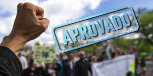 Imagem Acessível: A imagem mostra um punho erguido em primeiro plano, simbolizando conquista e resistência, com uma multidão desfocada ao fundo participando de uma manifestação em ambiente externo. No topo, há a hashtag “#DescongelaJá”, e sobre o céu azul aparece um grande carimbo azul com a palavra “APROVADO”. Na parte inferior, o texto diz: “Sancionada lei que garante direitos congelados na pandemia”. Na parte inferior central da imagem está o logotipo do SINJUS-MG.