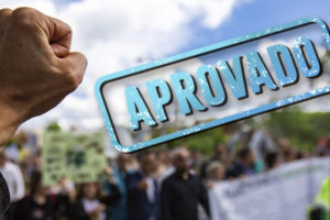 Imagem Acessível: A imagem mostra um punho erguido em primeiro plano, simbolizando conquista e resistência, com uma multidão desfocada ao fundo participando de uma manifestação em ambiente externo. No topo, há a hashtag “#DescongelaJá”, e sobre o céu azul aparece um grande carimbo azul com a palavra “APROVADO”. Na parte inferior, o texto diz: “Sancionada lei que garante direitos congelados na pandemia”. Na parte inferior central da imagem está o logotipo do SINJUS-MG.