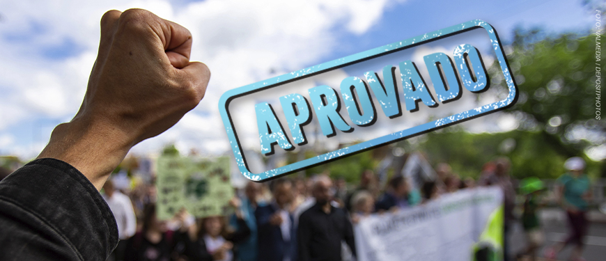 Imagem Acessível: A imagem mostra um punho erguido em primeiro plano, simbolizando conquista e resistência, com uma multidão desfocada ao fundo participando de uma manifestação em ambiente externo. No topo, há a hashtag “#DescongelaJá”, e sobre o céu azul aparece um grande carimbo azul com a palavra “APROVADO”. Na parte inferior, o texto diz: “Sancionada lei que garante direitos congelados na pandemia”. Na parte inferior central da imagem está o logotipo do SINJUS-MG.