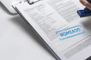 A imagem mostra uma mão segurando um carimbo azul e estampando a palavra "NOMEADO" em um documento preso a uma prancheta. O fundo contém itens de escritório, como um teclado, caneta e bloco de anotações. O texto sobreposto informa que, após intensa luta sindical, o TJMG nomeou os primeiros aprovados do concurso 01/2022, destacando essa conquista para os candidatos e sindicatos envolvidos.