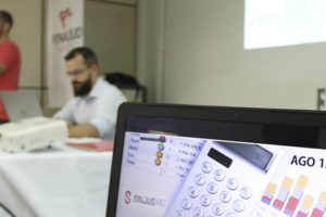 Em destaque está a tela de um computador com uma imagem de planilhas e calculadora, sobre elas há o título AGO - 13/04/2023. Ao fundo, estão o Coordenador-geral do Sindicato, Alexandre Pires e o diretor de Finanças, Felipe Rodrigues.
