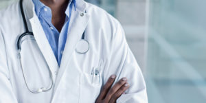 A imagem mostra ao fundo a foto do tronco de um profissional de saúde, vestindo jaleco branco com um estetoscópio pendurado no pescoço e os braços cruzados. No topo, lê-se “Déficit de servidores” e, em destaque ao centro, o texto diz: “Sindicatos reiteram solicitação de nomeação de médico para Polo de Saúde de Poços de Caldas”. Na parte inferior, aparece o símbolo do SINJUS.