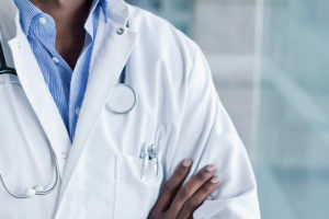 A imagem mostra ao fundo a foto do tronco de um profissional de saúde, vestindo jaleco branco com um estetoscópio pendurado no pescoço e os braços cruzados. No topo, lê-se “Déficit de servidores” e, em destaque ao centro, o texto diz: “Sindicatos reiteram solicitação de nomeação de médico para Polo de Saúde de Poços de Caldas”. Na parte inferior, aparece o símbolo do SINJUS.