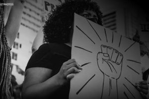 Foto em preto e branco de uma manifestação no qual mulheres e homens negros protestam contra o racismo. No centro da imagem está uma mulher negra, de máscara facial, segurando um cartaz com o desenho de um punho erguido.