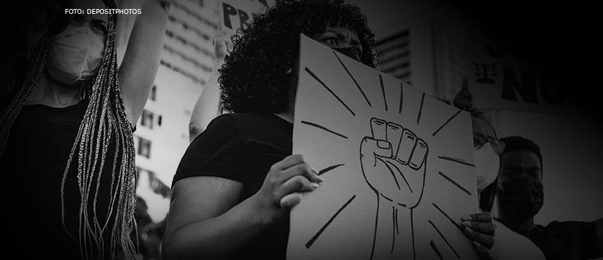 Foto em preto e branco de uma manifestação no qual mulheres e homens negros protestam contra o racismo. No centro da imagem está uma mulher negra, de máscara facial, segurando um cartaz com o desenho de um punho erguido.