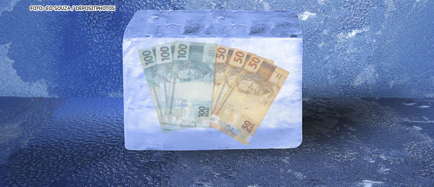 Imagem Acessível: Montagem digital com um conjunto de notas de 100 e 50 reais dentro de um cubo de gelo.