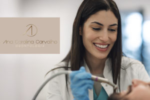 Imagem Acessível: Dentista jovem, de cabelos lisos e castanhos, sorri enquanto atende um paciente deitado na cadeira odontológica. Ela usa jaleco branco e luvas azuis. No canto esquerdo da imagem, há o logotipo “Ana Carolina Carvalho Odontologia”. Sobreposto à foto, está o texto: “Garanta saúde bucal e estética com o novo convênio do SINJUS”.