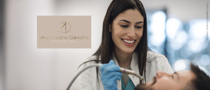 Imagem Acessível: Dentista jovem, de cabelos lisos e castanhos, sorri enquanto atende um paciente deitado na cadeira odontológica. Ela usa jaleco branco e luvas azuis. No canto esquerdo da imagem, há o logotipo “Ana Carolina Carvalho Odontologia”. Sobreposto à foto, está o texto: “Garanta saúde bucal e estética com o novo convênio do SINJUS”.
