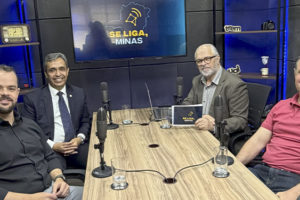Imagem acessível: A imagem mostra o registro fotográfico do último episódio do podcast Se Liga, Minas. Em destaque, o título diz: "Legado de JK atravessa gerações e inspira debate no Se Liga, Minas". Na base da imagem, aparece a logomarca do SINJUS-MG.