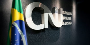 Imagem Acessível: Logotipo metálico do Conselho Nacional de Justiça (CNJ) em uma parede escura, parcialmente visível ao lado da bandeira do Brasil. Conteúdo textual: Articulação - SINJUS participa de audiência pública sobre os 20 anos do CNJ, nesta 3ªF., 14/10.