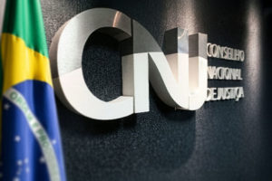 Imagem Acessível: Logotipo metálico do Conselho Nacional de Justiça (CNJ) em uma parede escura, parcialmente visível ao lado da bandeira do Brasil. Conteúdo textual: Articulação - SINJUS participa de audiência pública sobre os 20 anos do CNJ, nesta 3ªF., 14/10.