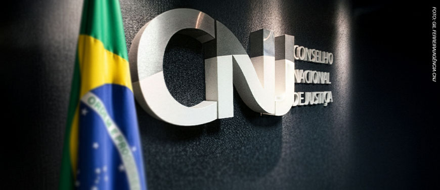Imagem Acessível: Logotipo metálico do Conselho Nacional de Justiça (CNJ) em uma parede escura, parcialmente visível ao lado da bandeira do Brasil. Conteúdo textual: Articulação - SINJUS participa de audiência pública sobre os 20 anos do CNJ, nesta 3ªF., 14/10.