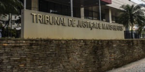 A imagem mostra o letreiro em frente ao edifício sede do Tribunal de Justiça de Minas Gerais (TJMG), as letras são de aço inox sobre um fundo de concreto bege. Atrás do letreiro é possível ver a entrada principal do Tribunal, uma construção revestida de vidro, onde há uma marquise com detalhe vermelho. Conteúdo textual: AVANÇO - Atuação sindical garante novas nomeações do Concurso 01/2022.