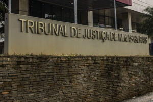 A imagem mostra o letreiro em frente ao edifício sede do Tribunal de Justiça de Minas Gerais (TJMG), as letras são de aço inox sobre um fundo de concreto bege. Atrás do letreiro é possível ver a entrada principal do Tribunal, uma construção revestida de vidro, onde há uma marquise com detalhe vermelho. Conteúdo textual: AVANÇO - Atuação sindical garante novas nomeações do Concurso 01/2022.
