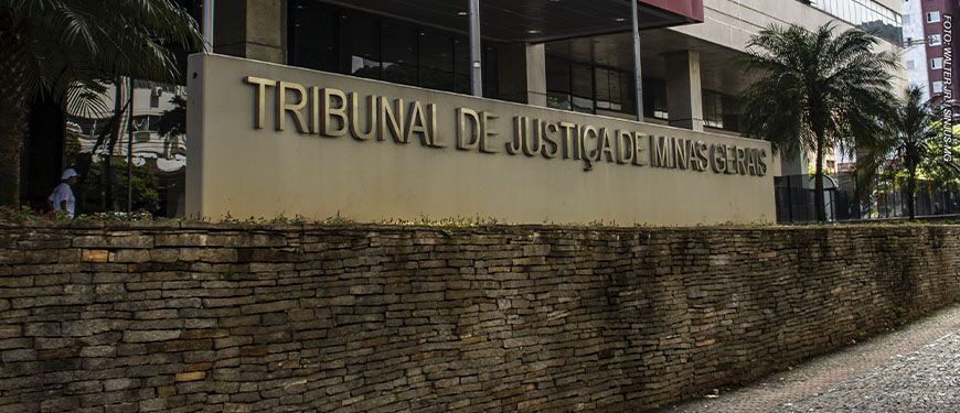A imagem mostra o letreiro em frente ao edifício sede do Tribunal de Justiça de Minas Gerais (TJMG), as letras são de aço inox sobre um fundo de concreto bege. Atrás do letreiro é possível ver a entrada principal do Tribunal, uma construção revestida de vidro, onde há uma marquise com detalhe vermelho. Conteúdo textual: AVANÇO - Atuação sindical garante novas nomeações do Concurso 01/2022.