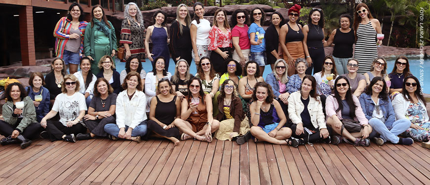 Foto de grupo do Núcleo das Mulheres do SINJUS-MG reunido em área externa, diante de uma piscina e cercado por vegetação tropical. As participantes, em sua maioria mulheres, posam sorridentes para a foto, expressando união e alegria. Conteúdo textual: CONFRADAY - Núcleo das Mulheres celebra ano de encontros e fortalecimento.