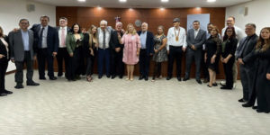 Grupo de pessoas integrantes de uma comitiva posam para foto em uma sala institucional durante visita oficial a autoridades do Paraguai. Todos estão alinhados em pé, formando uma fileira, vestidos de maneira formal, com painel de madeira e brasão ao fundo.