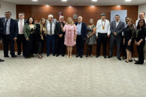Grupo de pessoas integrantes de uma comitiva posam para foto em uma sala institucional durante visita oficial a autoridades do Paraguai. Todos estão alinhados em pé, formando uma fileira, vestidos de maneira formal, com painel de madeira e brasão ao fundo.