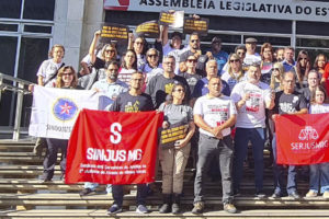 Grupo de representantes do Serjusmig, SINDOJUS/MG e SINJUS-MG, reunidos para foto em frente à entrada da ALMG, eles carregam bandeiras dos três sindicatos e cartazes em protesto contra o RRF do Governo Zema (Regime de Recuperação Fiscal). Entre os sindicalistas está o diretor de Imprensa e Comunicação do SINJUS-MG, Alexandre Gomes.